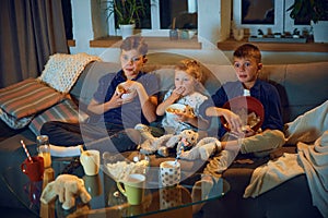 Children, brothers and little sister sitting on couch in living room in evening, watching tv together and eating snacks.