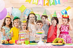 Children celebrating birthday party. Kids stand at festive table and show thumbs up