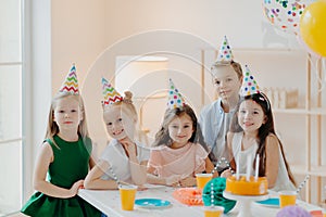 Children, celebration and birthday concept. Positive children have fun together at party, wear cone hats, eat delicious cake, play