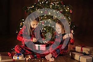 Children in checkered red pajamas are unpacking Christmas presents sitting on floor at night by Xmas tree.