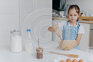 Children, cooking and home concept. Pretty dark haired girl whisks ingredients in bowl, busy preparing dough for cake, being