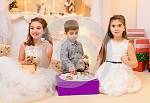 Children drink tea, eat cookies and play with the toy dishes in home interior decorated with holiday lights and gifts