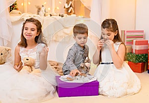 Children drink tea, eat cookies and play with the toy dishes in home interior decorated with holiday lights and gifts