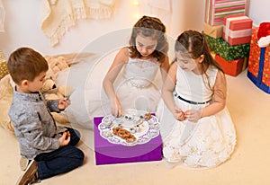 Children drink tea, eat cookies and play with the toy dishes in home interior decorated with holiday lights and gifts