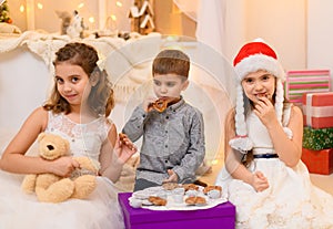 Children drink tea, eat cookies and play with the toy dishes in home interior decorated with holiday lights and gifts