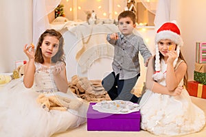 Children drink tea, eat cookies and play with the toy dishes in home interior decorated with holiday lights and gifts