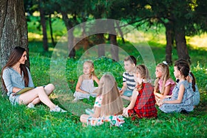 Children and education, young woman at work as educator reading book to boys and girls in park.