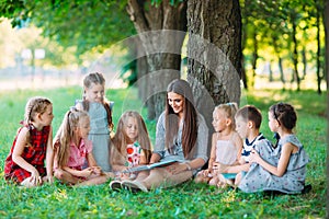 Children and education, young woman at work as educator reading book to boys and girls in park.