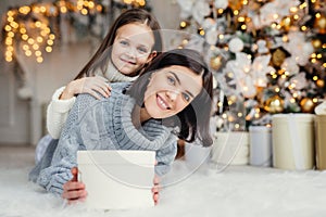 Children, family and celebration concept. Adorable female in knitted sweater holds white present box and small kid stands behind