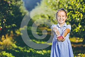 Cute little girl stands over blurred autumn garden background, extending big red apple in her palms to the camera