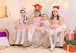 Children girls portrait, sisters dressed in santa helper hat, sitting on a couch in home interior decorated with christmas lights