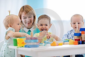 Children group playing with teacher in day care centre. Carer and babies at table in nursery playroom