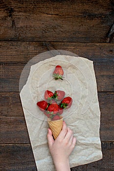 Children hand takes strawberries in ice cream cone on natural wooden background. Strawberries in wafel cone and Baby girl hand.