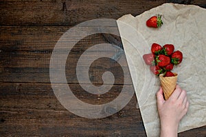 Children hand takes strawberries in ice cream cone on natural wooden background. Strawberries in wafel cone and Baby girl hand.