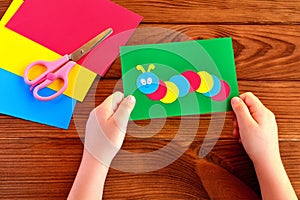 Children hands holding paper applique caterpillar. Paper sheets, scissors on a brown wooden background