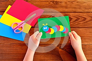 Children hands holding paper applique caterpillar. Paper sheets, scissors on a brown wooden background