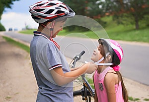 Children learn to ride bicycle in a park on summer day. Teenager boy helping preschooler girl to put on safety helmet