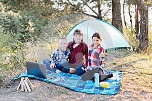 Children looking show on laptop in the forest. Boys and girls are resting in a tent camp. Children sit on the plaid on the grass