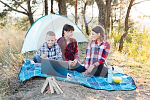 Children looking show on laptop in the forest. Boys and girls are resting in a tent camp. Children sit on the plaid on the grass