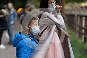 Children in masks look at animals through fence. Boy and girl wearing warm clothes walking in zoo in autumn. Quarantine