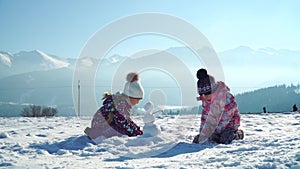 Children in outwear making small snowman while playing on snowy field in sunlight with mountains on background