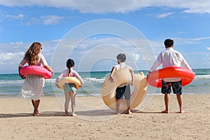 Children with parent wearing colorful inflatable swim ring running to blue sea, kids and adult in a line from behind on tropical