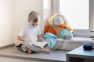 Children play a doctor. A large brown toy in a medical bandage and shoe covers is sitting on the window. A child is reading a book