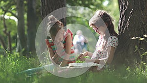 Children read a book outdoors sitting on the grass near a tree. Two little girls have fun together