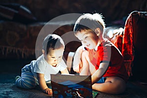 Children reading a book sitting together on floor at home. brother and babysister smiling having fun with book together. Boy and