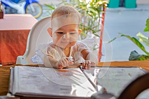 Children  reading chooses and studies the menu in the restaurant. Infant girl is sitting on a baby`s high chair in a street cafe