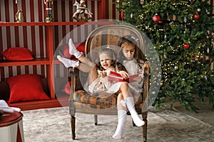 Children are reading Christmas book, sitting in chair by the Xmas tree