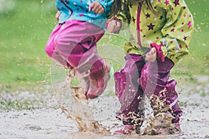 Children in rubber boots and rain clothes jumping in puddle.