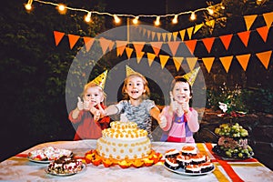 Children`s birthday party. Three cheerful children girls at the table eating cake with their hands and smearing their face. Fun a