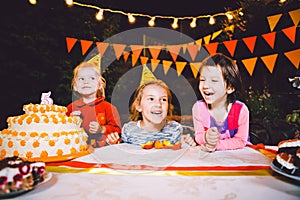 Children`s birthday party. Three cheerful children girls at the table eating cake with their hands and smearing their face. Fun a