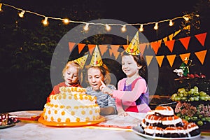 Children`s birthday party. Three cheerful children girls at the table eating cake with their hands and smearing their face. Fun a