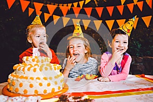 Children`s birthday party. Three cheerful children girls at the table eating cake with their hands and smearing their face. Fun a