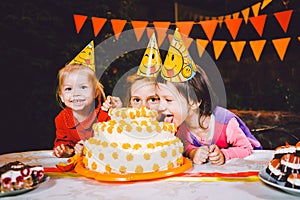 Children`s birthday party. Three cheerful children girls at the table eating cake with their hands and smearing their face. Fun a