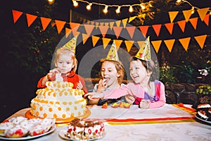 Children`s birthday party. Three cheerful children girls at the table eating cake with their hands and smearing their face. Fun a