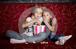 Children`s cinema: A girl and a boy watch a movie at home on a big red sofa in the dark and eat popcorn.