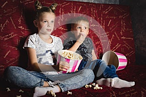 Children`s cinema: A girl and a boy watch a movie at home on a big red sofa in the dark and eat popcorn.