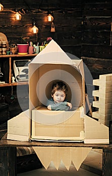 Children`s dreams of space. Kid sit in cardboard hand made rocket. Boy play at home with rocket, little cosmonaut sit in