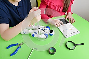 Children's hands assemble robot on workbench, screwing parts while reviewing programming code on tablet