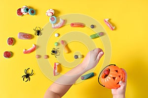 Children's hands collect marmalades for Halloween with a pumpkin-shaped basket on a yellow background with a place