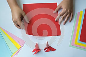 Children`s hands do origami butterfly from colored paper on white background.