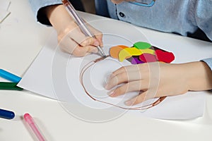 Children`s hands hold a brown felt-tip pen and paint on a white piece of paper.