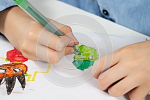 Children`s hands hold a green felt-tip pen and draw a green guy on a white piece of paper.