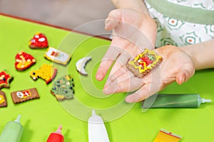 Children`s hands are holding a decorated rectangular cake. Colorful cinnamon cakes lie on the green table in the background. Tubes