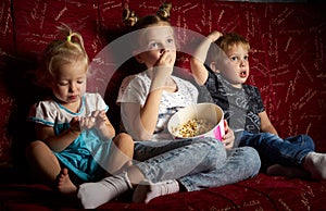 Children`s movies: Three children watch movies at home on a big red sofa in the dark and eat popcorn.