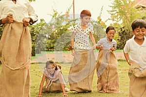 Children`s playing potato sack jumping race at park outdoor - kids falling and having fun while playing gunny sack race.