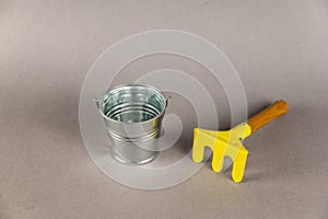 Children`s toys on a gray background. A small empty metal bucket and a yellow rake with a wooden handle. Garden tools
